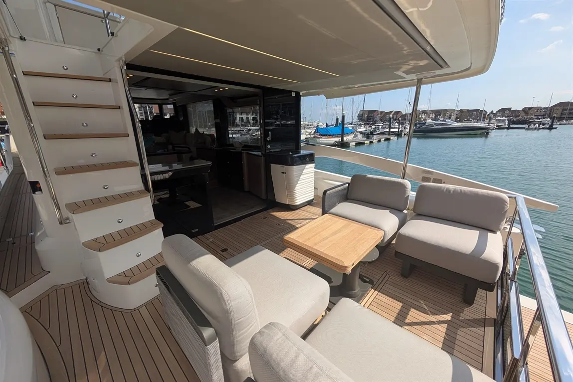 Luxury yacht deck featuring grey seating, wooden table, and stairs leading to interior, with boats visible in the marina.